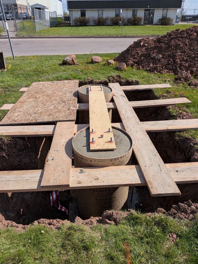 Concrete support piers under construction with wooden framing and formwork, surrounded by excavated dirt and industrial buildings in background.